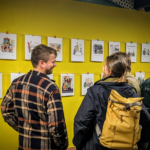 A group of people laughing (hopefully at the cartoons) at two rows of cartoons hung across a yellow wall at an exhibition.