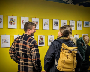 A group of people laughing (hopefully at the cartoons) at two rows of cartoons hung across a yellow wall at an exhibition.
